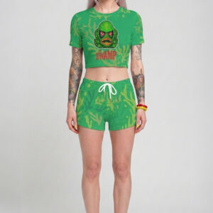Mujer con cabello lavanda pastel posando con un conjunto verde de dos piezas estilo gótico pastel, que incluye un crop top de manga corta con un monstruo verde retro que dice 'The Swamp' y unos shorts cortos a juego con cordón blanco.