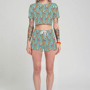 A model with pastel lavender hair wearing a pink two-piece pastel goth outfit, featuring a short-sleeve crop top and matching shorts with a seamless pattern of melting eyeball ice cream cones.