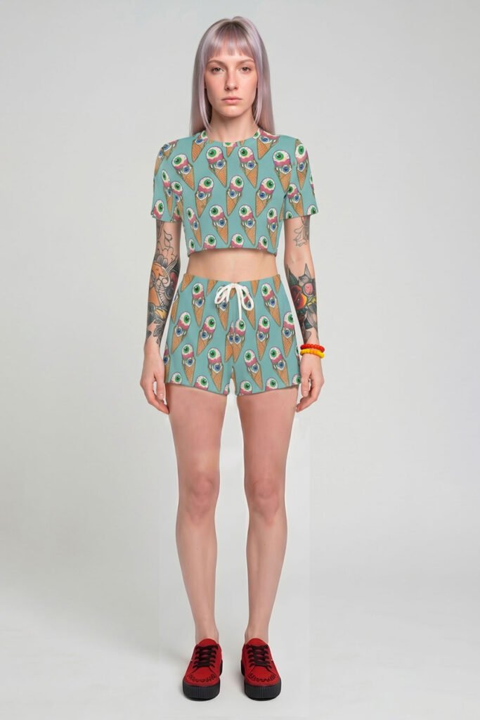 A model with pastel lavender hair wearing a pink two-piece pastel goth outfit, featuring a short-sleeve crop top and matching shorts with a seamless pattern of melting eyeball ice cream cones.