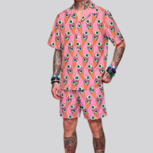 A tattooed male model with slicked-back blonde hair wearing a pink two-piece pastel goth outfit, featuring a short-sleeve button-down shirt and matching shorts with a seamless pattern of melting eyeball ice cream cones.
