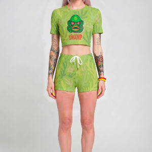 A model with pastel lavender hair wearing a two-piece pastel goth green outfit, featuring a short-sleeve crop top with a retro green monster graphic that says 'The Swamp' and matching shorts with a white drawstring