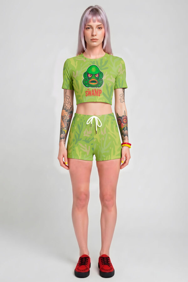 A model with pastel lavender hair wearing a two-piece pastel goth green outfit, featuring a short-sleeve crop top with a retro green monster graphic that says 'The Swamp' and matching shorts with a white drawstring